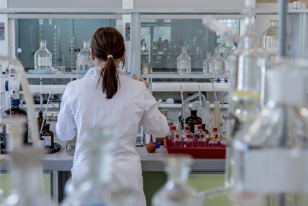 woman in lab coat with back to camera, in a laboratory with test tubes and equipment in foreground and background.